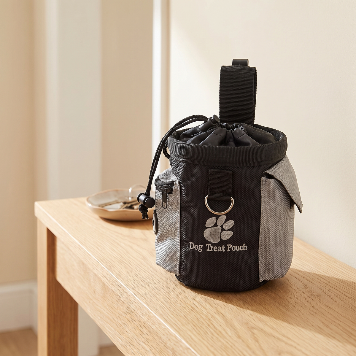 Black and grey dog treat pouch with drawstring top and belt clip standing on an oak console table