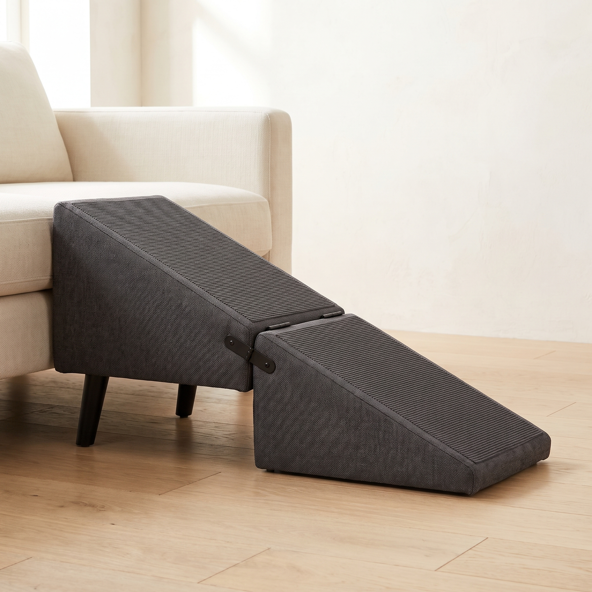 Charcoal grey folding dog ramp opened in front of a low cream sofa, on black tapered legs